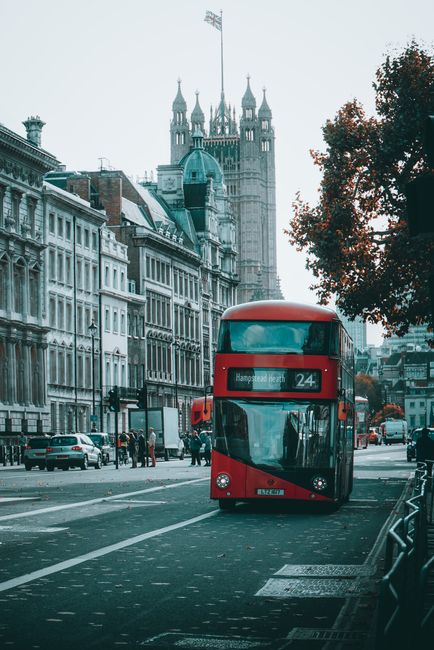 A red double-decker bus numbered 24 is traveling along a street in London, with the Houses of Parliament and the Big Ben clock tower visible in the background. The bus displays its destination as Hamp